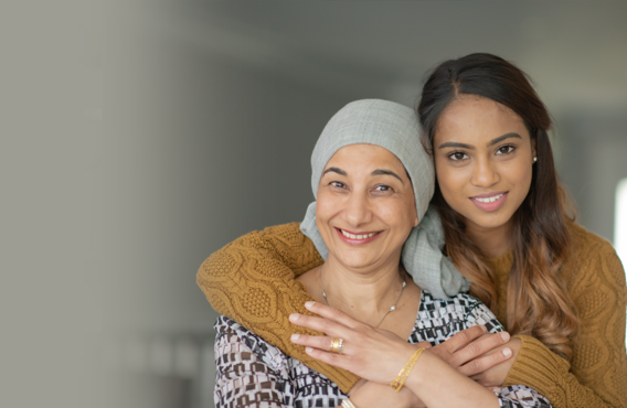 Une jeune femme serre dans ses bras une femme plus âgée par derrière. Tous deux sourient et la femme plus âgée a un tissu enroulé sur la tête.