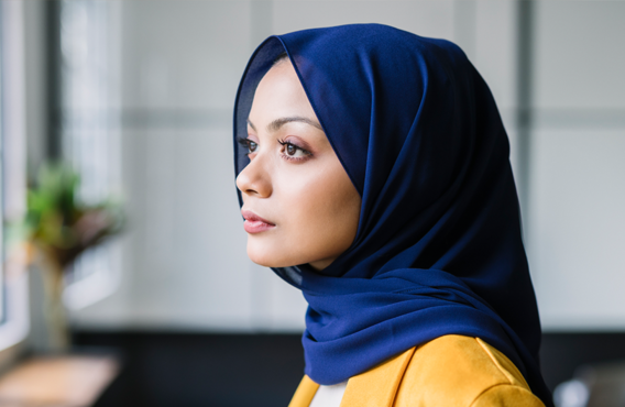 Femme portant un hijab bleu debout près d'une fenêtre donnant sur l'extérieur.