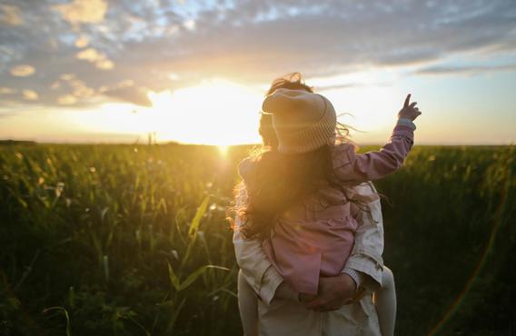 A woman carrying a little girl in a field of corn in front of a bright evening sun