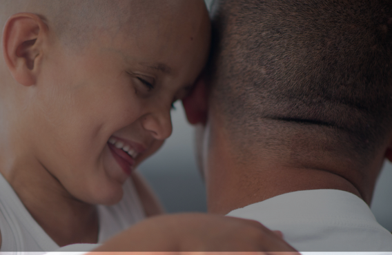 Child with a shaved head embraces an adult, with faces close together in a bright indoor setting.