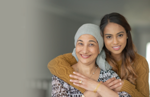 Une jeune femme serre dans ses bras une femme plus âgée par derrière. Tous deux sourient et la femme plus âgée a un tissu enroulé sur la tête.