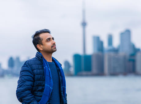 Un homme debout près d’un port de Toronto, regardant au loin, les mains dans les poches. Derrière lui se trouve le paysage du centre-ville de Toronto avec la tour CN visible.