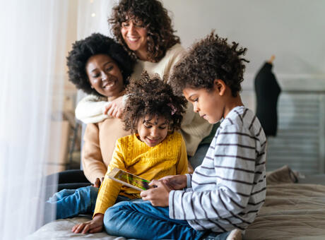 une famille de 4 personnes assises sur un lit. Les deux parents s'embrassent et les deux enfants jouent avec une tablette.