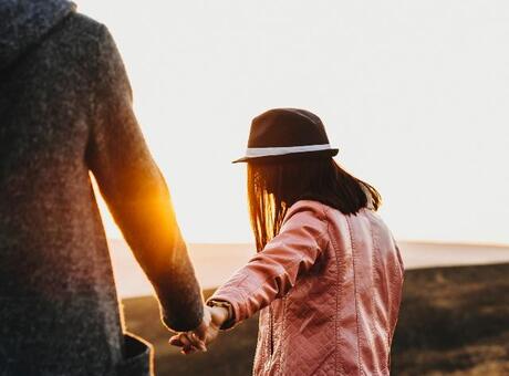 Une personne vêtue d'une veste rose et coiffée d'un chapeau traverse un paysage ouvert en tenant la main d'une autre personne. La lumière chaude du coucher de soleil brille entre elles, créant un éclat lumineux qui se détache sur le décor naturel.