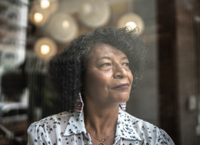 An older Black woman with colourful earrings looks out a window.