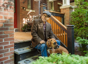 A man dressed in a tweed jacket and hat sits on a stoop. His dog is with him.