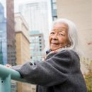 A woman with long white hair stands outside on a balcony in a city and looks at the camera