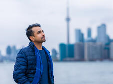 Un homme debout près d’un port de Toronto, regardant au loin, les mains dans les poches. Derrière lui se trouve le paysage du centre-ville de Toronto avec la tour CN visible.