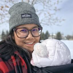 A woman stands outside in a snowy landscape. She has a young child in a baby carrier on her chest.