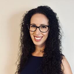 A White woman with long curly hair and glasses smiles at the camera.