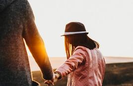 Une personne vêtue d'une veste rose et coiffée d'un chapeau traverse un paysage ouvert en tenant la main d'une autre personne. La lumière chaude du coucher de soleil brille entre elles, créant un éclat lumineux qui se détache sur le décor naturel.